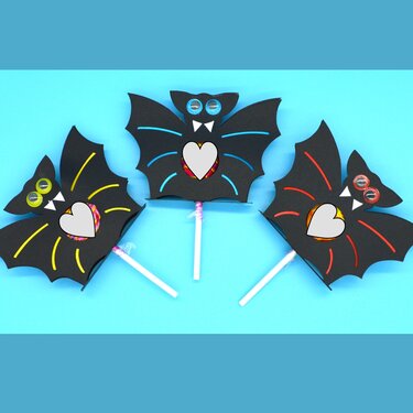 Drei schwarze Papierfledermäuse mit Wackelaugen, die jeweils einen Lutscher in einer herzförmigen Aussparung halten, auf blauem Hintergrund.