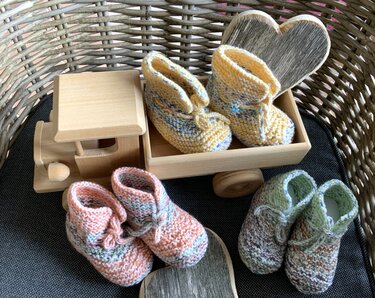 Drei Paare handgestrickter Babyschuhe in Pastellgelb, Pfirsich und Hellgrün mit Bindebändchen, arrangiert im Weidenkorb neben Holzspielzeug und Holzherzen.