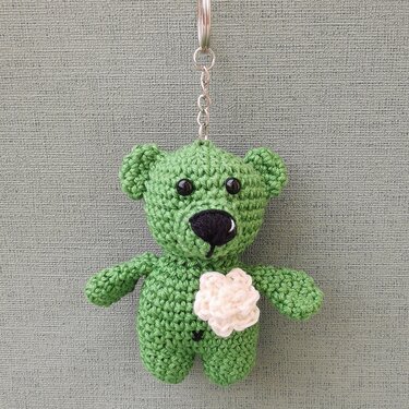 kleiner grüner gehäkelter Teddybär-Schlüsselanhänger mit cremefarbener Blüte vor grauem Hintergrund