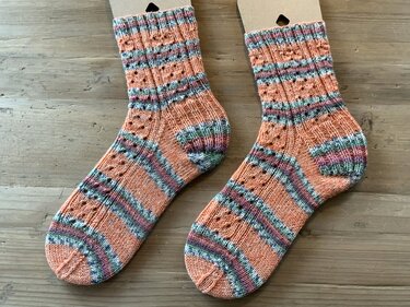Paar kurzschaftiger handgestrickter Socken in Apricot mit Rippbündchen, Ajourlochmuster und bunten Streifen auf Holzuntergrund
