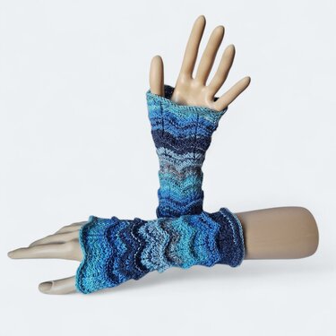 Zwei Mannequin‑Hände tragen gestrickte, fingerlose Armstulpen in Blautönen mit Wellenmuster vor weißem Hintergrund.