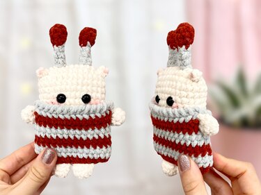 Zwei kleine gehäkelte Amigurumi mit cremefarbenem Kopf, schwarzen Sicherheitsaugen und rot-grau gestreiftem Körper, in Händen gehalten