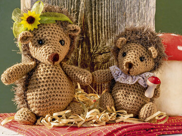 Zwei gehäkelte Amigurumi-Igel mit Blume, Spitzenkragen und kleinem Pilz vor Holzhintergrund