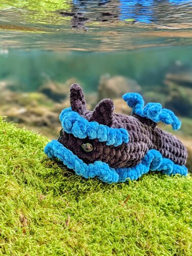 Braun-blaue gehäkelte Amigurumi-Meeresschnecke auf grünem Moos vor Wasser