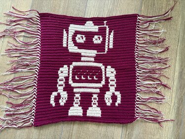 Handgehäkeltes Rechteck mit weißem Pixel‑Roboter auf magentafarbenem Hintergrund und Fransen