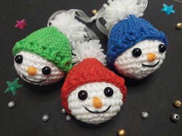Drei gehäkelte Schneemann-Anhänger mit Mützen in Grün, Rot und Blau auf dunklem Hintergrund mit Sternen und Perlen.