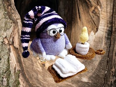 Gehäkelte lila Amigurumi-Eule mit gestreifter Schlafmütze, runder Brille, gehäkelter Kerze und gefalteten weißen Stoffstücken in einer Baumhöhle