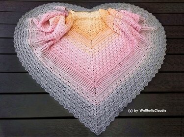 Herzförmiges Häkeltuch mit Pastellverlauf (Gelb, Rosa, Grau) auf dunkler Holzfläche