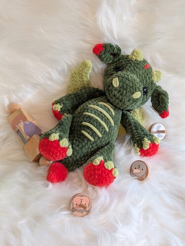 Handgearbeitetes dunkelgrünes Amigurumi‑Drachenplüschtier mit roten Akzenten und hellgrünen Details, liegend auf weißem Flauschteppich