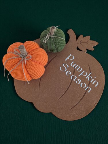 Braunes Kürbisschild mit weißer Aufschrift 'Pumpkin Season' und zwei gestrickten Kürbissen auf dunkelgrünem Hintergrund