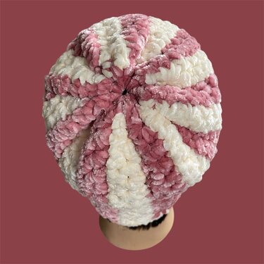 Draufsicht auf eine gehäkelte Beanie-Mütze mit rosa und cremefarbenen, radiär angeordneten Streifen und plüschiger Textur.
