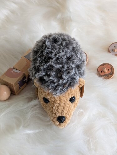 Gehäkelter Amigurumi-Igel mit beigem Gesicht und grauem, flauschigem Rücken, liegend auf hellem Untergrund