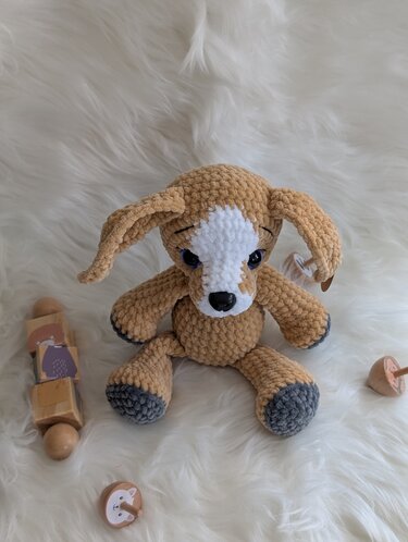 Beiger gehäkelter Amigurumi-Hund mit weißem Gesichtsfleck und grauen Pfoten, sitzend auf flauschigem weißem Untergrund