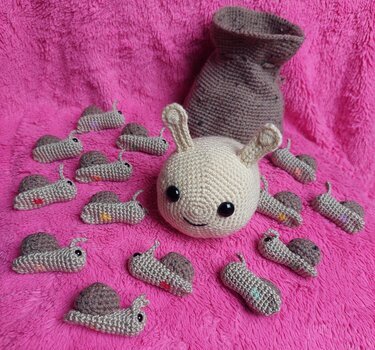 Große, helle Amigurumi-Figur mit Ohren umgeben von vielen kleinen gehäkelten Schnecken auf pinkem Plüschstoff.