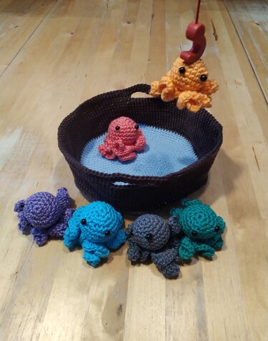 Handgehäkelte Kraken in verschiedenen Farben, Korb mit blauem Innenboden und Angel auf hellem Holzboden