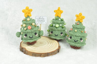 Drei gehäkelte Mini-Weihnachtsbäume mit gelben Sternen und Perlen, einer auf einer Holzscheibe mit 'Merry Christmas'-Schild