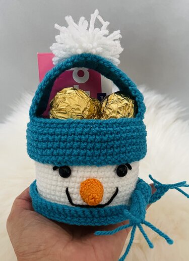 Hand hält gehäkelten Schneemannkorb in Weiß und Türkis mit Bommelmütze, Schal und goldenen Süßigkeiten