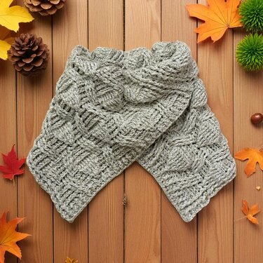 Grey crocheted scarf with basketweave texture on wooden planks with autumn leaves and pinecones.