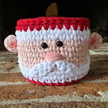 Runder, gehäkelter Korb mit Weihnachtsmann-Gesicht, roter Krempe und weißem Bart auf Ziegelboden.