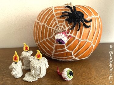 Orangefarbener Kürbis mit weißem Garn-Spinnennetz, schwarzer gehäkelter Spinne, gehäkelten Augäpfeln und kleinen geschmolzenen Kerzen auf Holztisch.