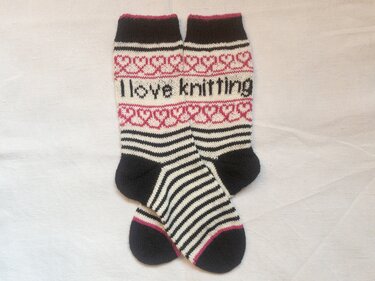 Unisex‑Socken in Schwarz, Creme und Pink mit Streifen, Herzbordüre und dem Schriftzug ‚I love knitting‘.