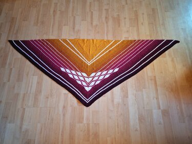 Gestricktes Dreieckstuch mit Farbverlauf von Gelb über Orange und Magenta zu Dunkelrot, weiße Streifen und zentrales Zopfmuster auf hellem Holzboden.