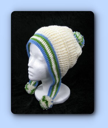 Cremefarbene gehäkelte Mütze mit blau-grünen Streifen an den Ohrenklappen und drei Bommeln auf einer Schaufensterpuppe