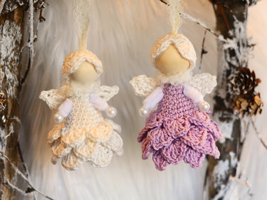 Zwei gehäkelte Amigurumi-Engel, einer cremefarben und einer in hellem Lila, mit kleinen Flügeln und Perlen, aufgehängt an winterlichen Zweigen vor hellem Hintergrund.