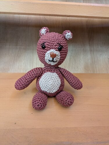 Gehäkelter Amigurumi-Teddybär in Altrosa mit hellem Bauch und Schnauze, sitzend auf Holz