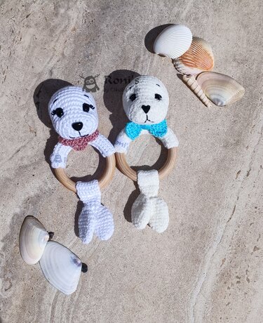 Zwei gehäkelte Seehund-Beißringe mit Holzringen auf Sand neben Muscheln
