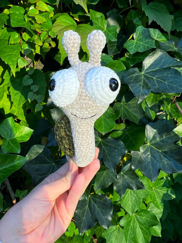 Handgehäkelte Amigurumi-Schnecke, beiger Körper, grünes Schneckenhaus, große Augen vor Efeu