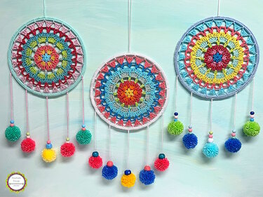 Three crocheted dreamcatchers with mandala centers, decorative beads, and colorful pom-poms on a pale blue background.