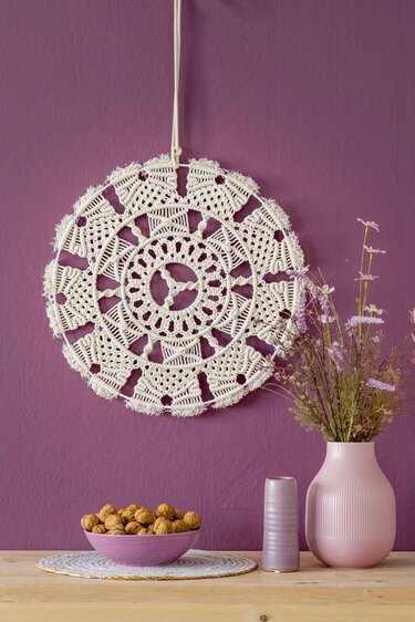 Runder cremefarbener Makramee-Wandbehang mit radialem Muster vor violetter Wand, neben Vase und Schale mit Nüssen.