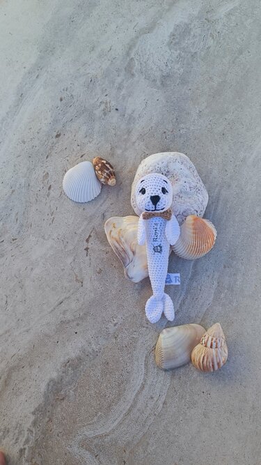 Weiß gehäkelte Seehund-Rassel mit brauner Schleife auf Muscheln im Sand