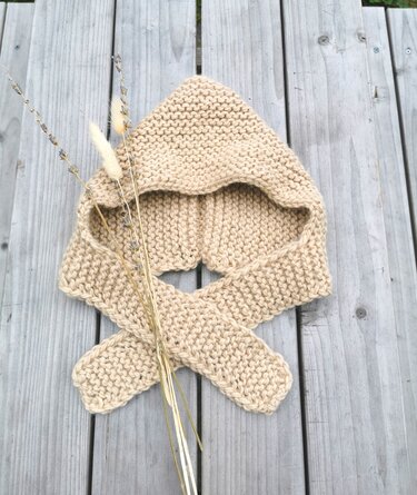 Beigefarbene gestrickte Babykapuze mit gekreuzten Schalenden auf grauem Holztisch, dekoriert mit getrockneten Gräsern.