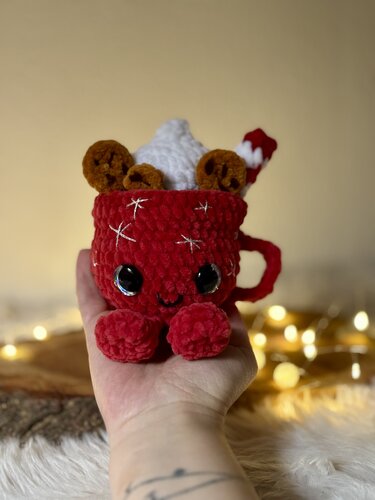 Rote gehäkelte Amigurumi-Tasse mit Gesicht, gehäkelter Sahne, Keksen und Zuckerstange.