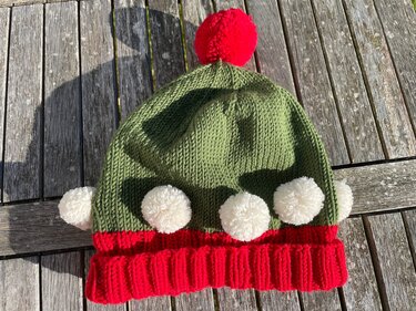 Gestrickte grün-rote Mütze mit rotem Bommel oben und weißen Bommeln am Saum auf Holzbrett