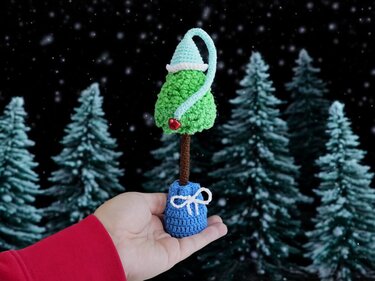 Hand hält kleinen grünen gehäkelten Weihnachtsbaum mit blauer Zipfelmütze vor verschneitem Waldhintergrund