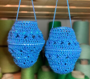 Zwei blaue gehäkelte Lampions mit Perlen und Lochmuster, hängend vor Garnspulen.