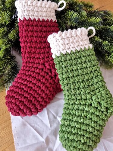 Zwei gehäkelte Weihnachtsstrümpfe in Rot und Grün mit cremefarbenen Bündchen und Aufhängeschlaufen vor Tannengrün.