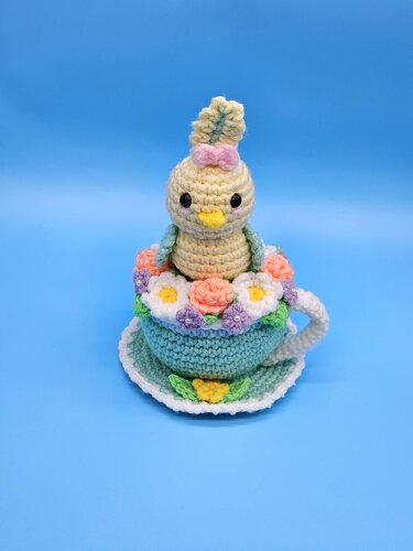 Crocheted turquoise teacup and saucer filled with pastel flowers and topped with a small chick figure on a blue background.