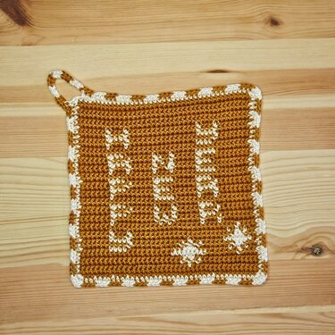 Quadratischer gehäkelter Topflappen in Senfgelb mit weißem Schriftzug „LEBEN LIEBEN LACHEN“, Sternmotiven und weißer Bordüre, auf Holz liegend.