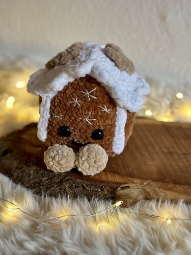 Gehäkeltes Lebkuchenhäuschen-Amigurumi mit weißem Zuckerguss, schwarzen Augen und Pompon-Füßen auf Holzscheibe vor Lichterkette