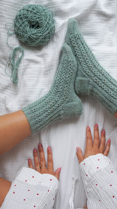 Gestrickte mintgrüne Socken mit Lochmuster und Garnkugel auf weißer Bettwäsche, sichtbare Hände im Vordergrund