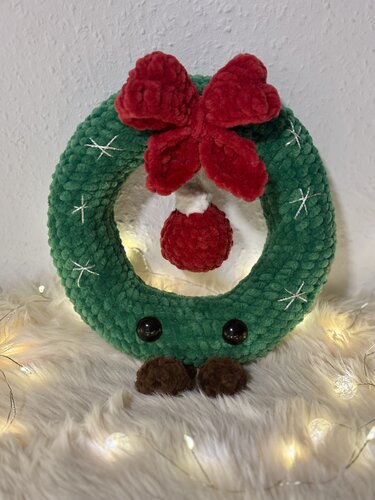 Grüner gehäkelter Weihnachtskranz mit roter Schleife, rotem Anhänger, stilisierten Augen und braunen Füßen auf hellem, flauschigem Untergrund mit Lichterketten.