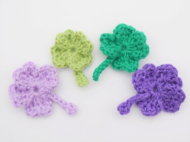 Vier gehäkelte Kleeblätter in hellgrün, grasgrün, hellviolett und violett auf hellem Untergrund