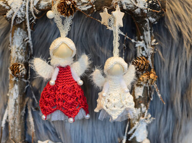 Zwei gehäkelte Engel-Anhänger, einer rot, einer weiß, mit Perlen und Tüll an Spitzenbändern vor winterlichem Kranz