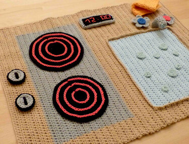 Gehäkelter Spielteppich in Herdform mit Kochfeldern, Bedienknöpfen, Digitalanzeige und Spülbereich.