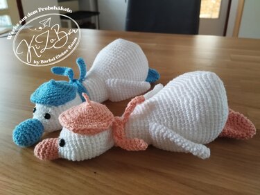 Nahaufnahme von drei weißen Amigurumi-Enten auf einem Holztisch; zwei tragen hellblaue bzw. pfirsichfarbene Mützen.