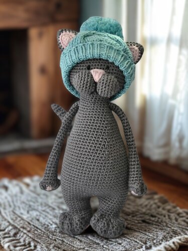 Gehäkelte graue Amigurumi‑Katze mit hellblauer Bommelmütze, stehend auf einem Teppich